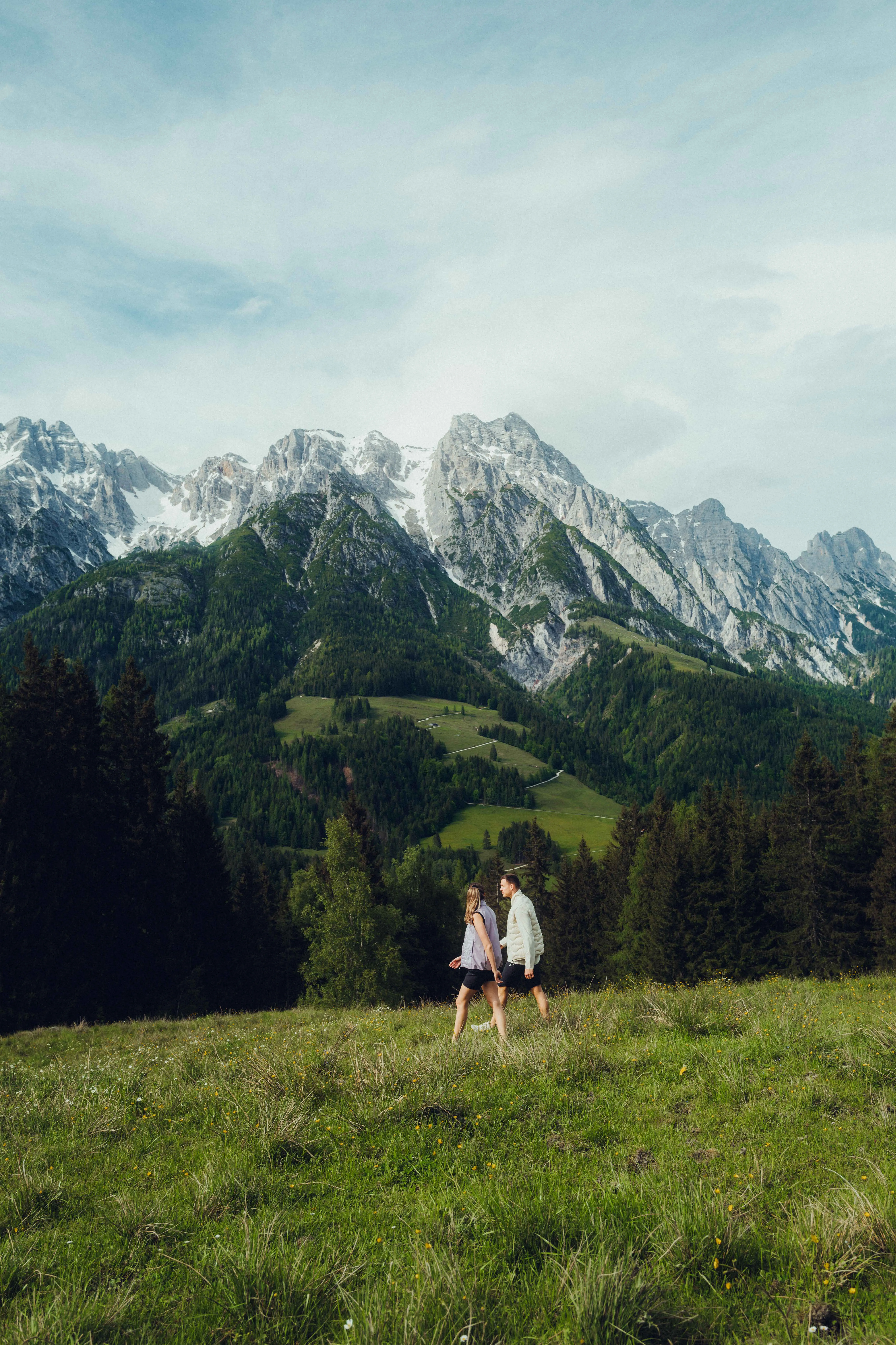 Zwei Personen gehen durch eine Wiese mit Bergen im Hintergrund.