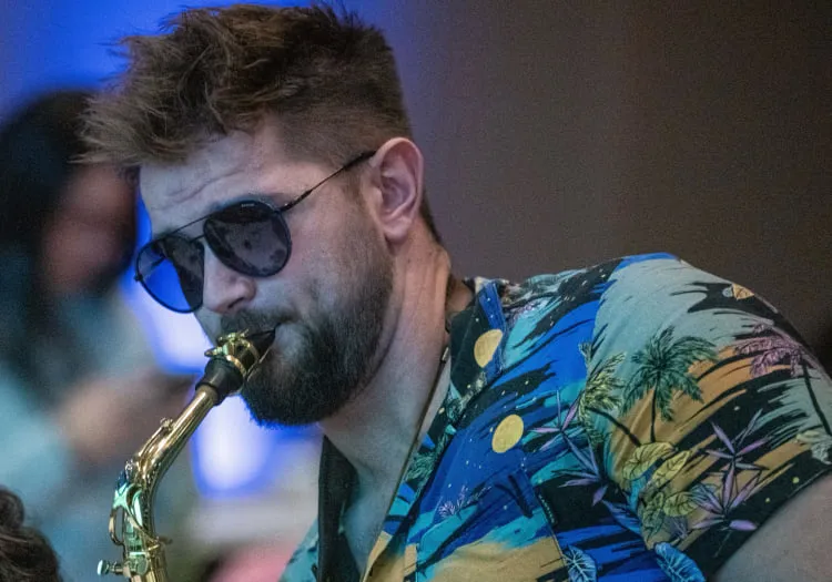 Man in sunglasses plays saxophone, colorful palm-patterned shirt