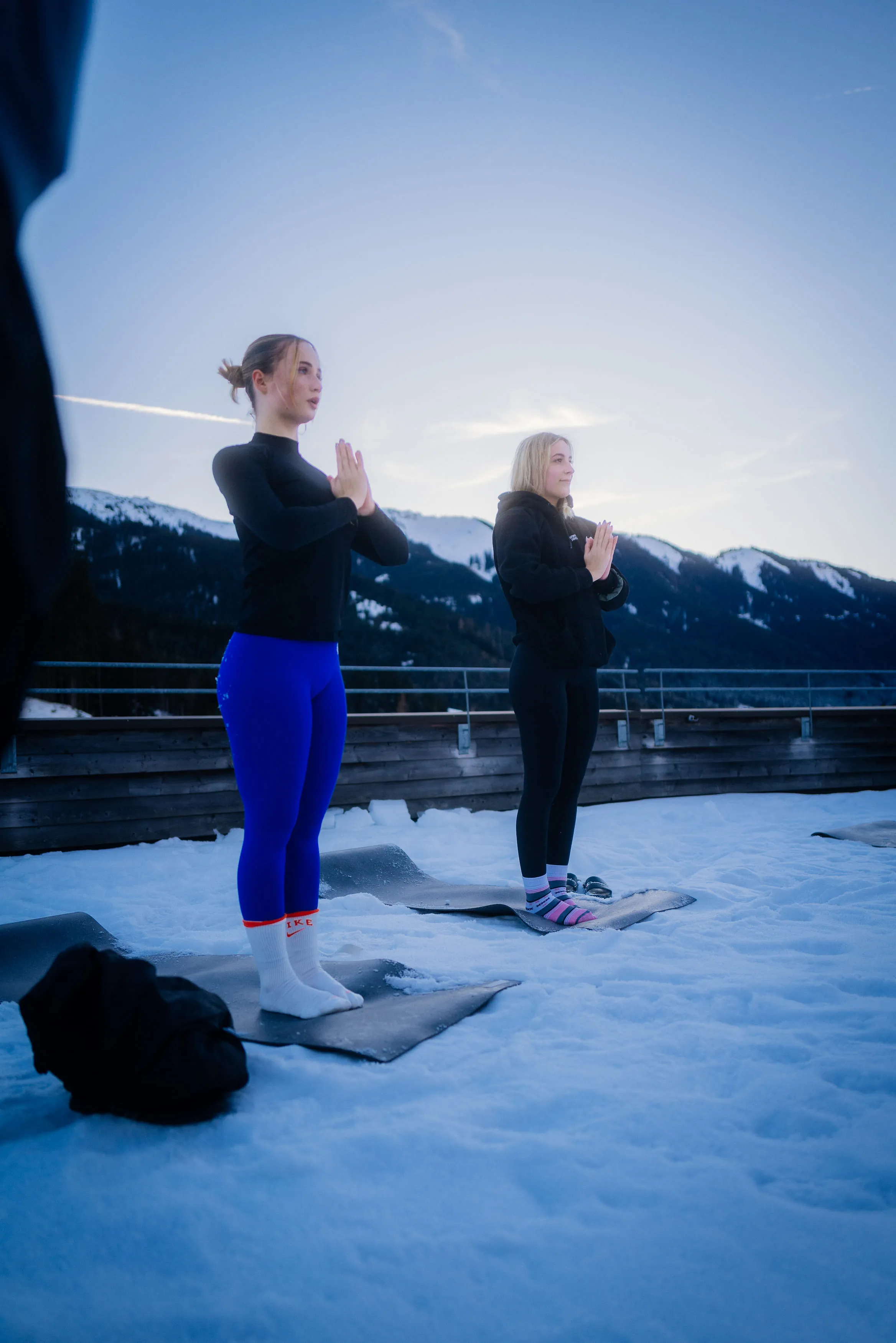 Zwei Personen praktizieren Yoga auf Matten im Schnee mit Bergen im Hintergrund