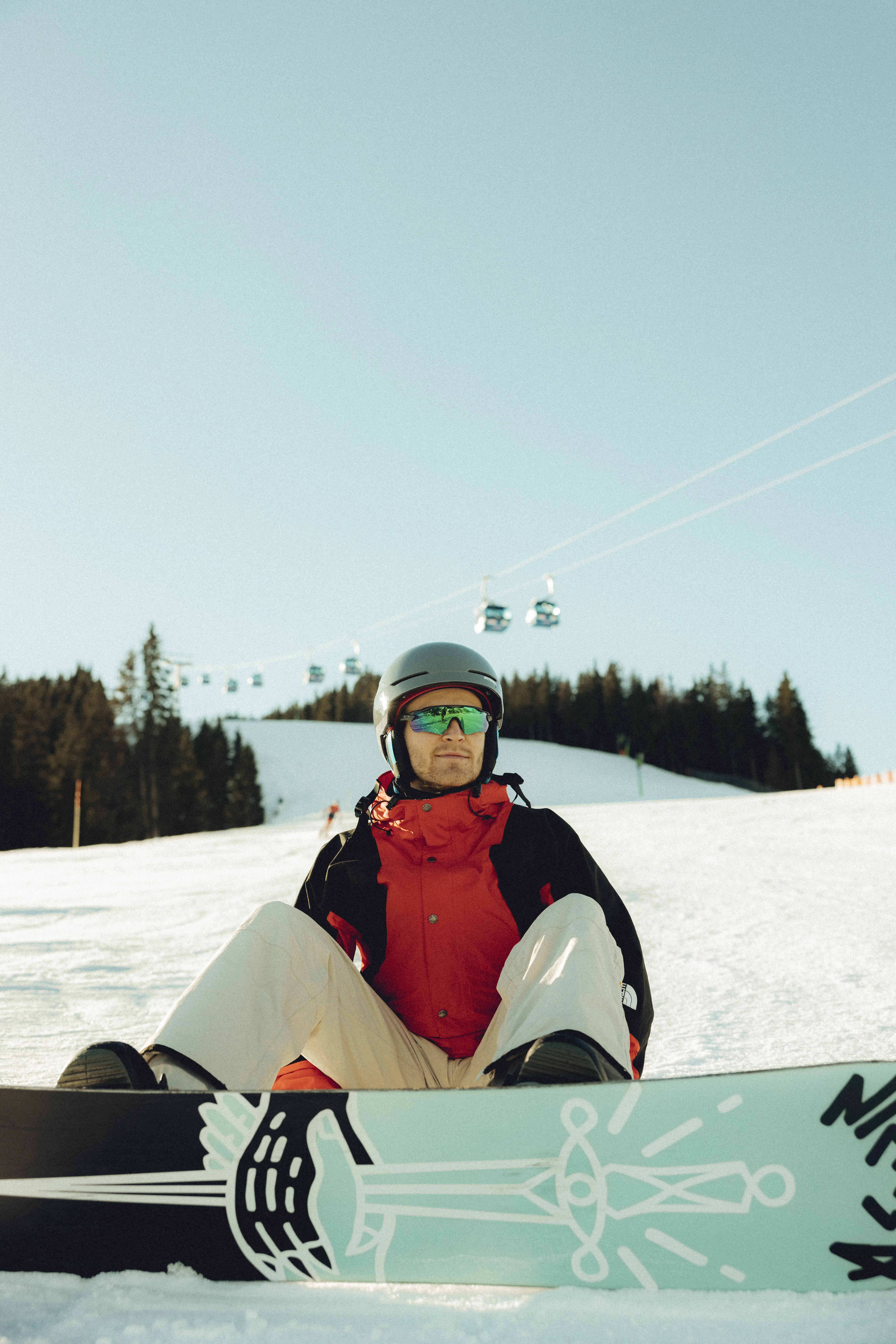 Snowboarder sitzt auf der Piste bei Forsthofalm, Seilbahnen im Hintergrund