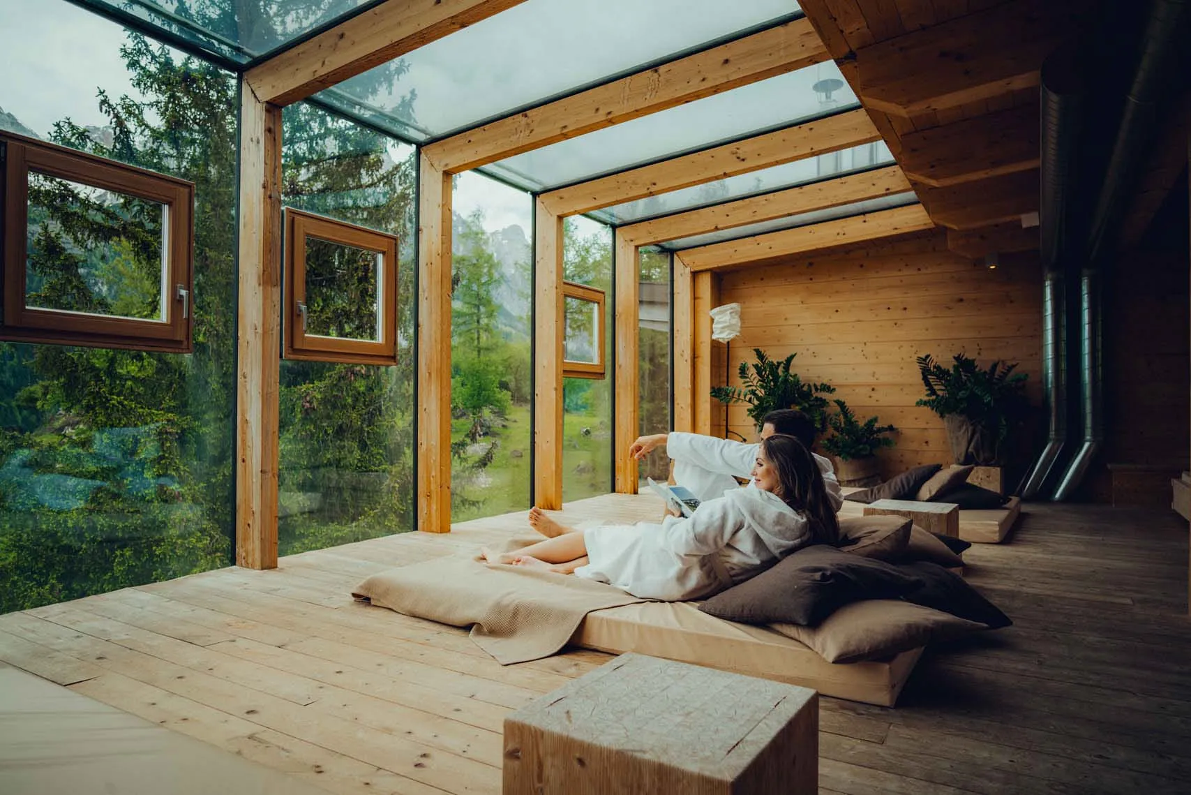 Modern wooden structure with large windows and wooden decor, person relaxing on cushions.