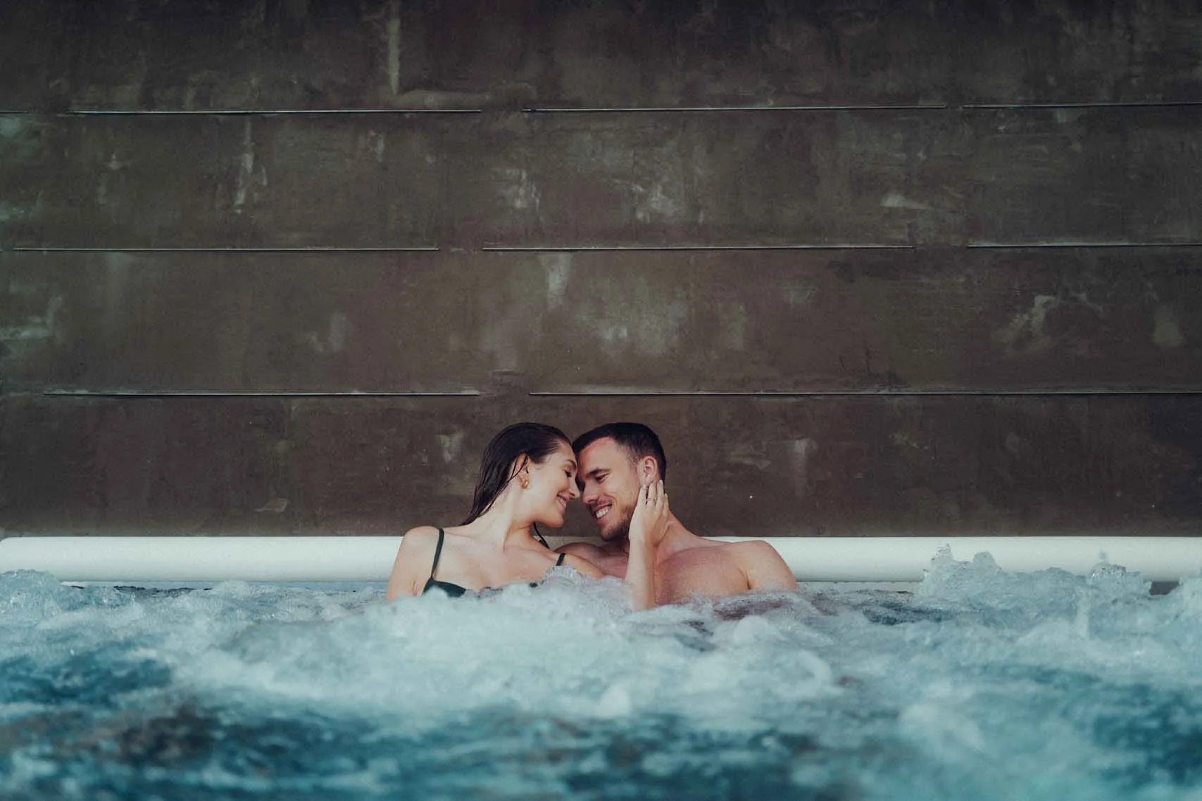 Couple in a pool, smiling and gazing at each other, against a gray wall.