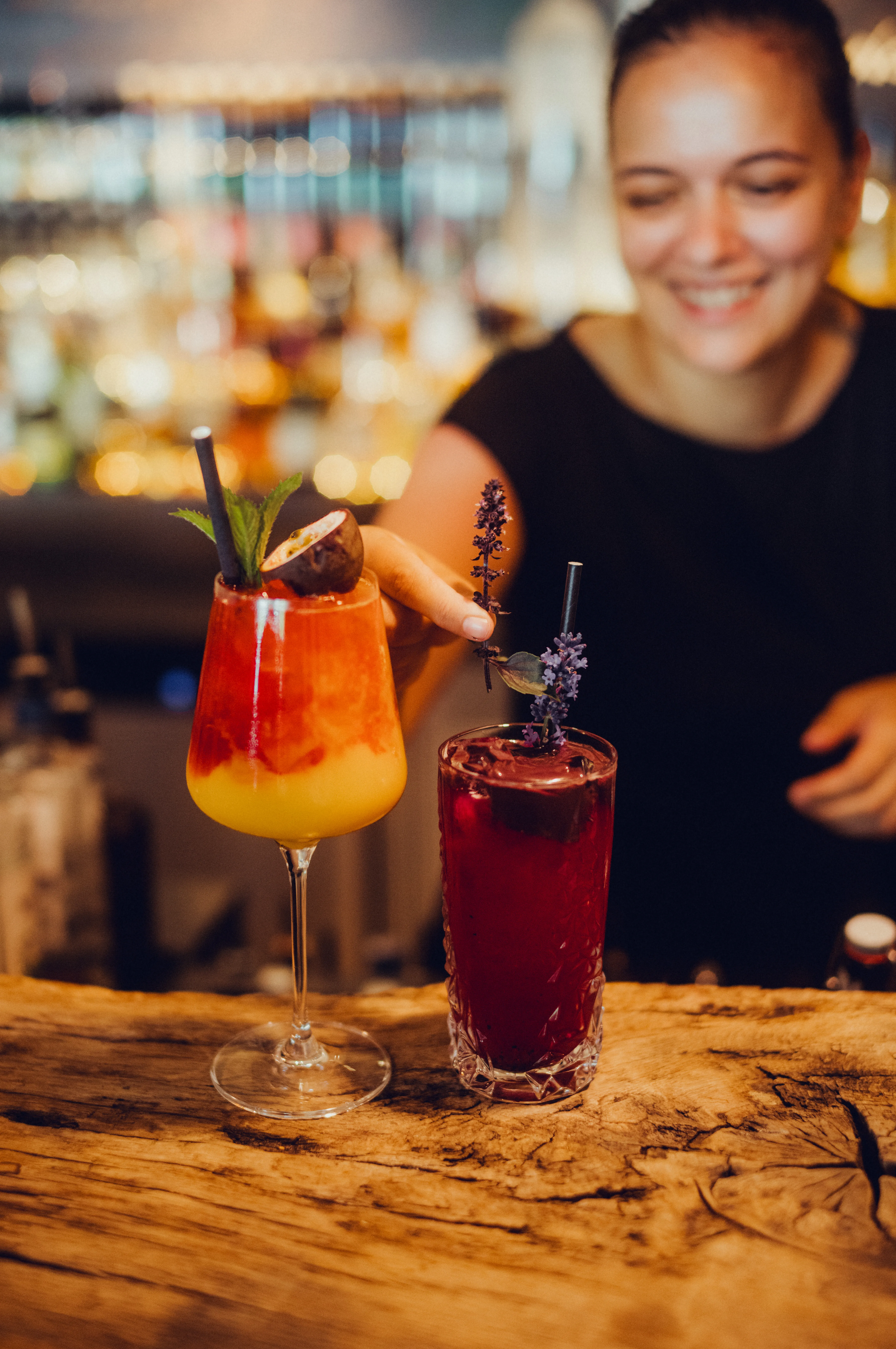 Two colorful cocktails on a wooden bar with a smiling bartender in the background
