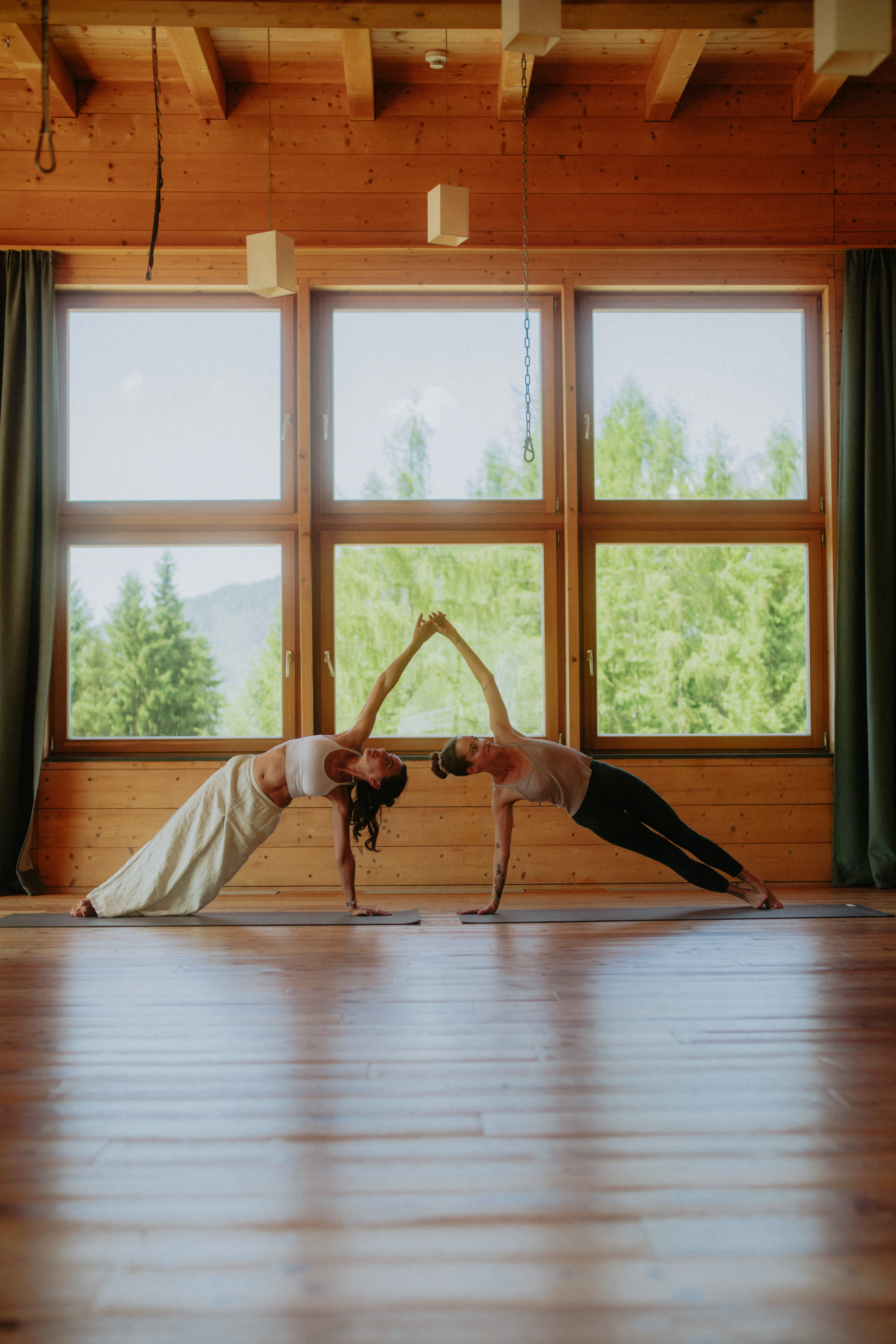 Two women in yoga poses in a wooden room with windows and green view