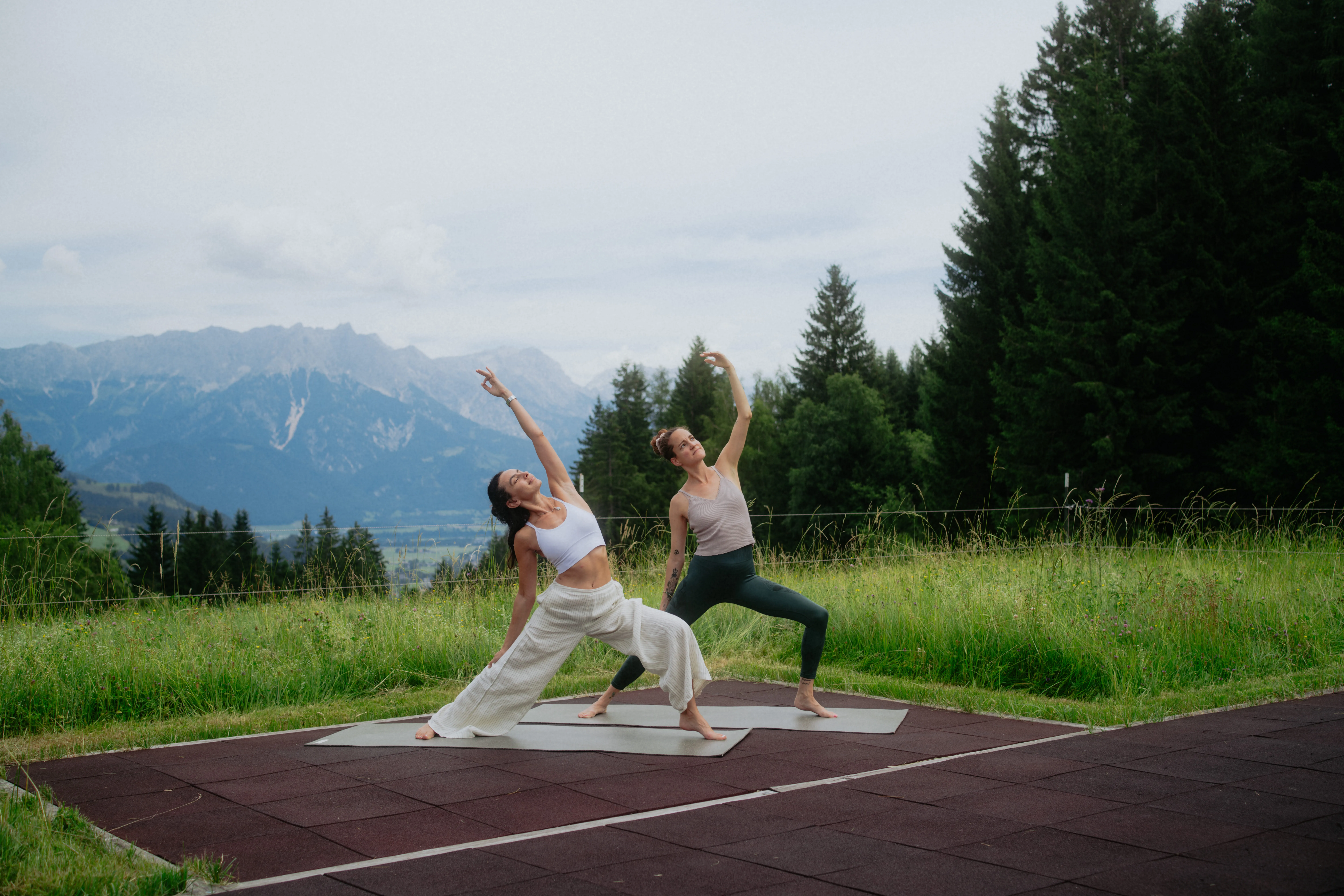 Two women practicing yoga outdoors on mats with mountains in the background