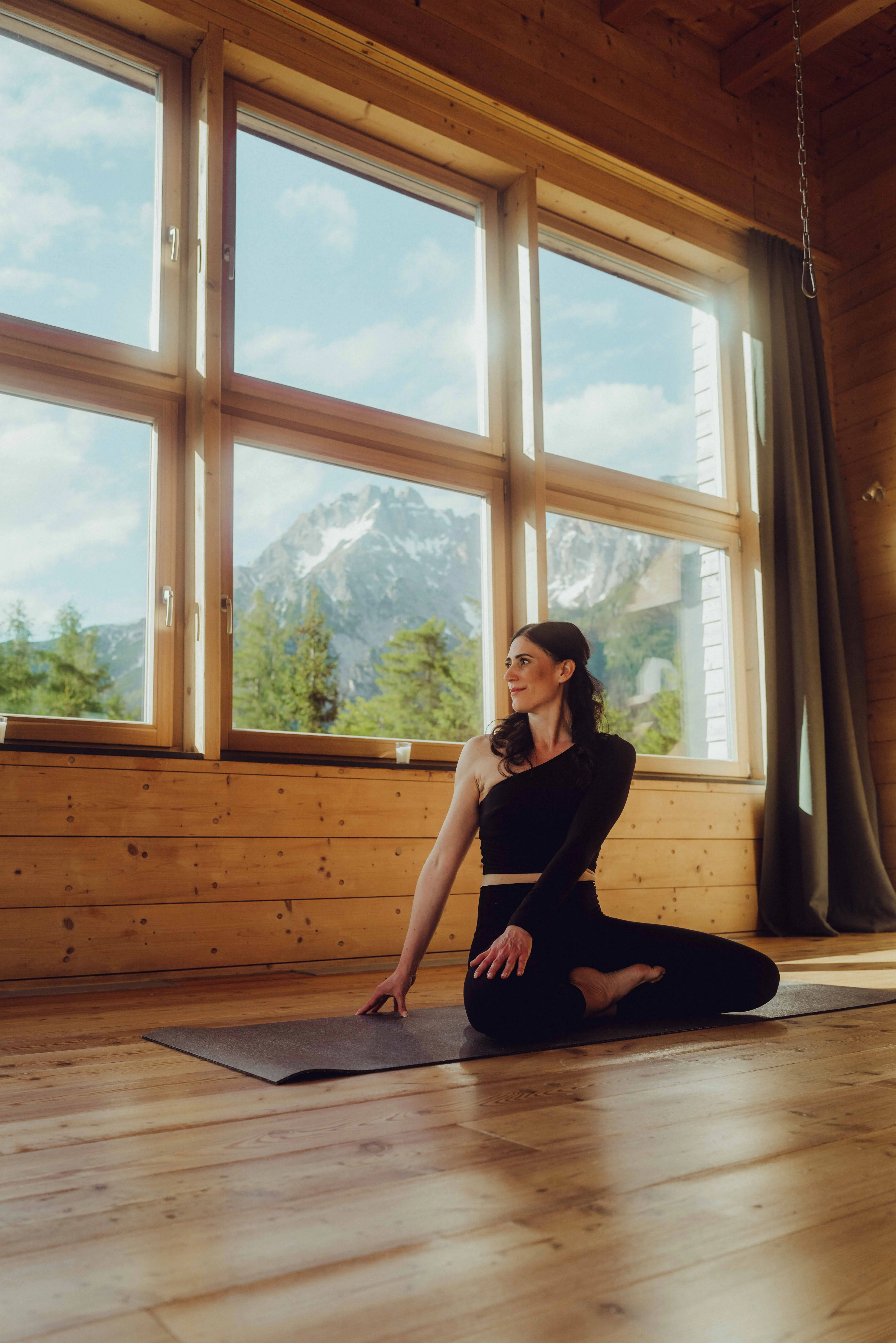 Woman in yoga pose with mountain view behind large windows in wooden cabin.