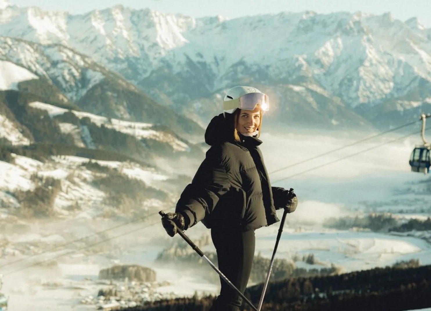 Person lächelt auf einem schneebedeckten Berg mit Skilift und Bergen im Hintergrund