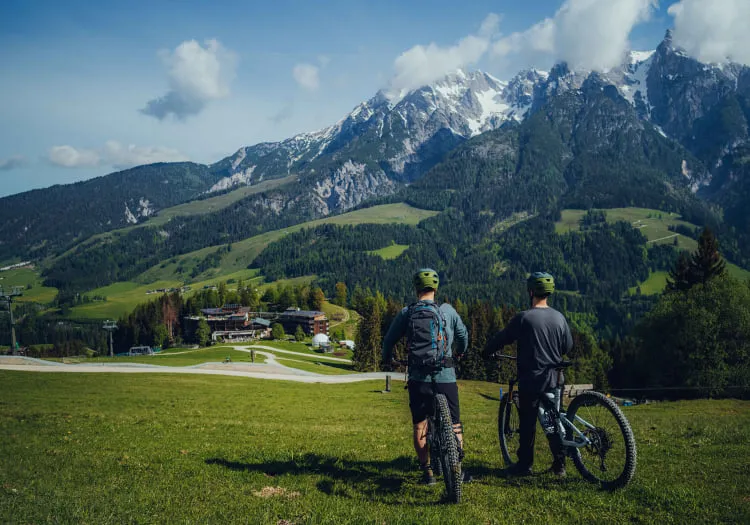 Zwei Mountainbiker stehen auf einer Wiese vor einer Berglandschaft.