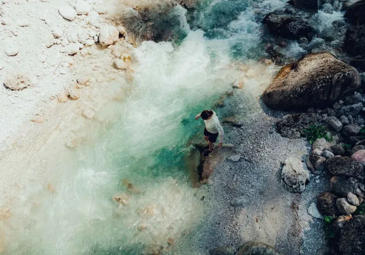 Person steht am Ufer eines klaren Flusses mit Steinen.