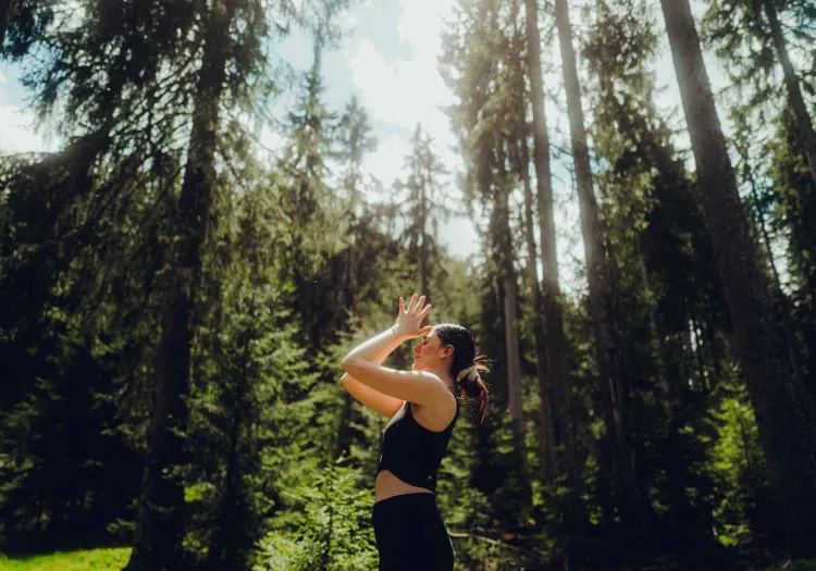 Frau im Wald mit erhobenen Händen, umgeben von hohen Bäumen.