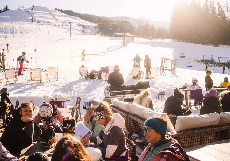People on a terrace in the snow with a ski area in the background.