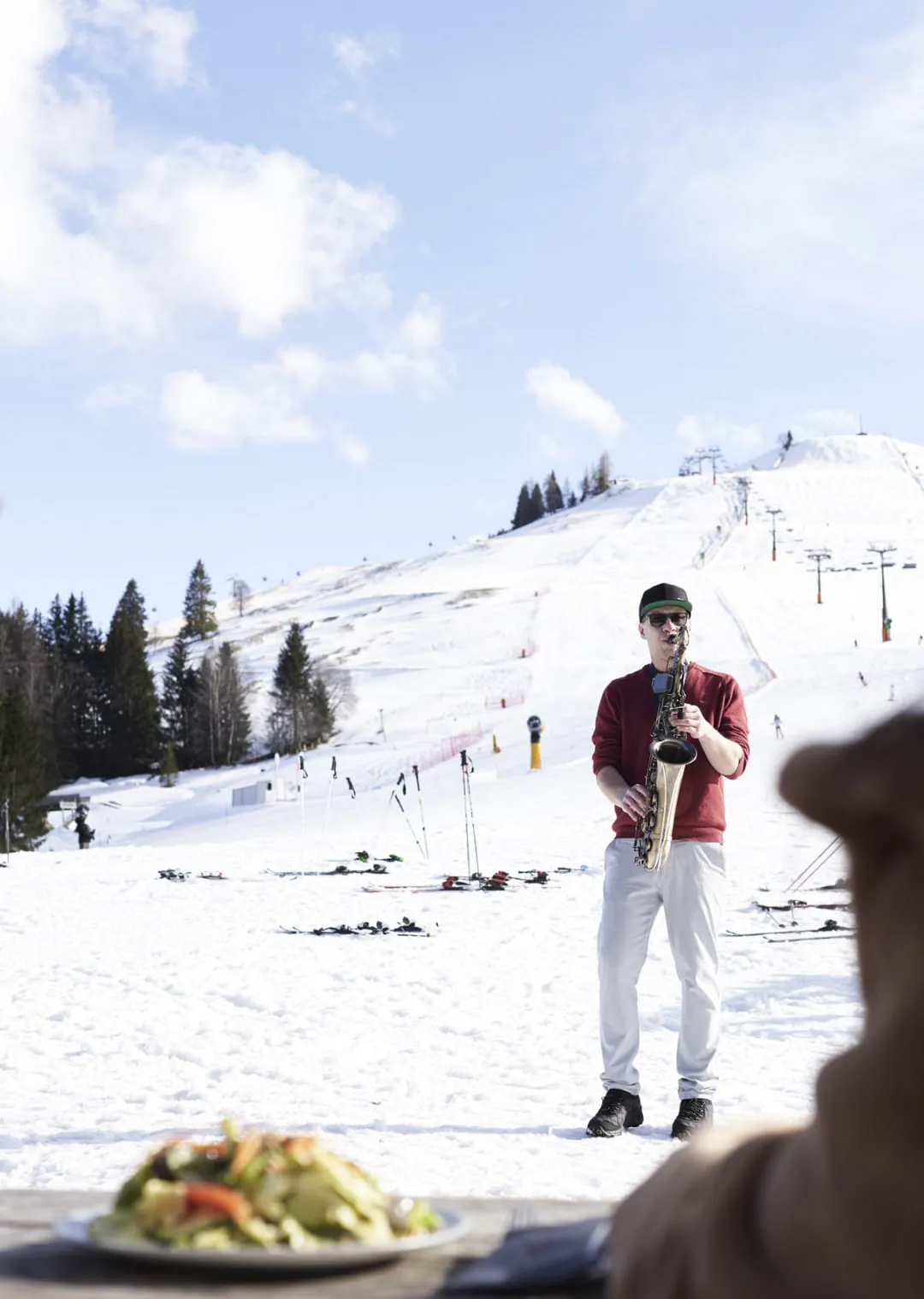 Mann spielt Saxophon vor schneebedecktem Hang, Winterlandschaft mit Skipisten.