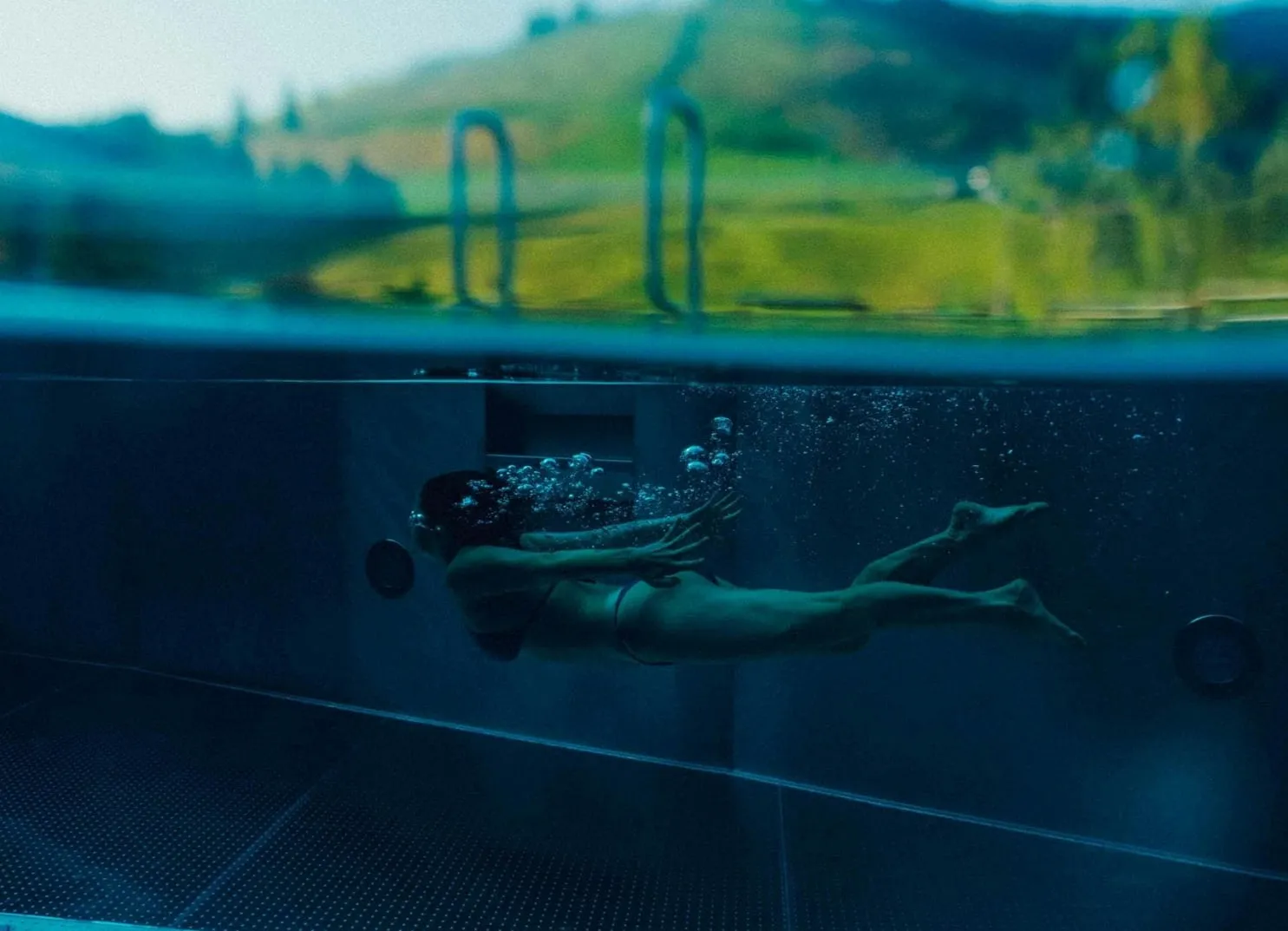 A person swimming underwater in a pool with a view of the landscape.