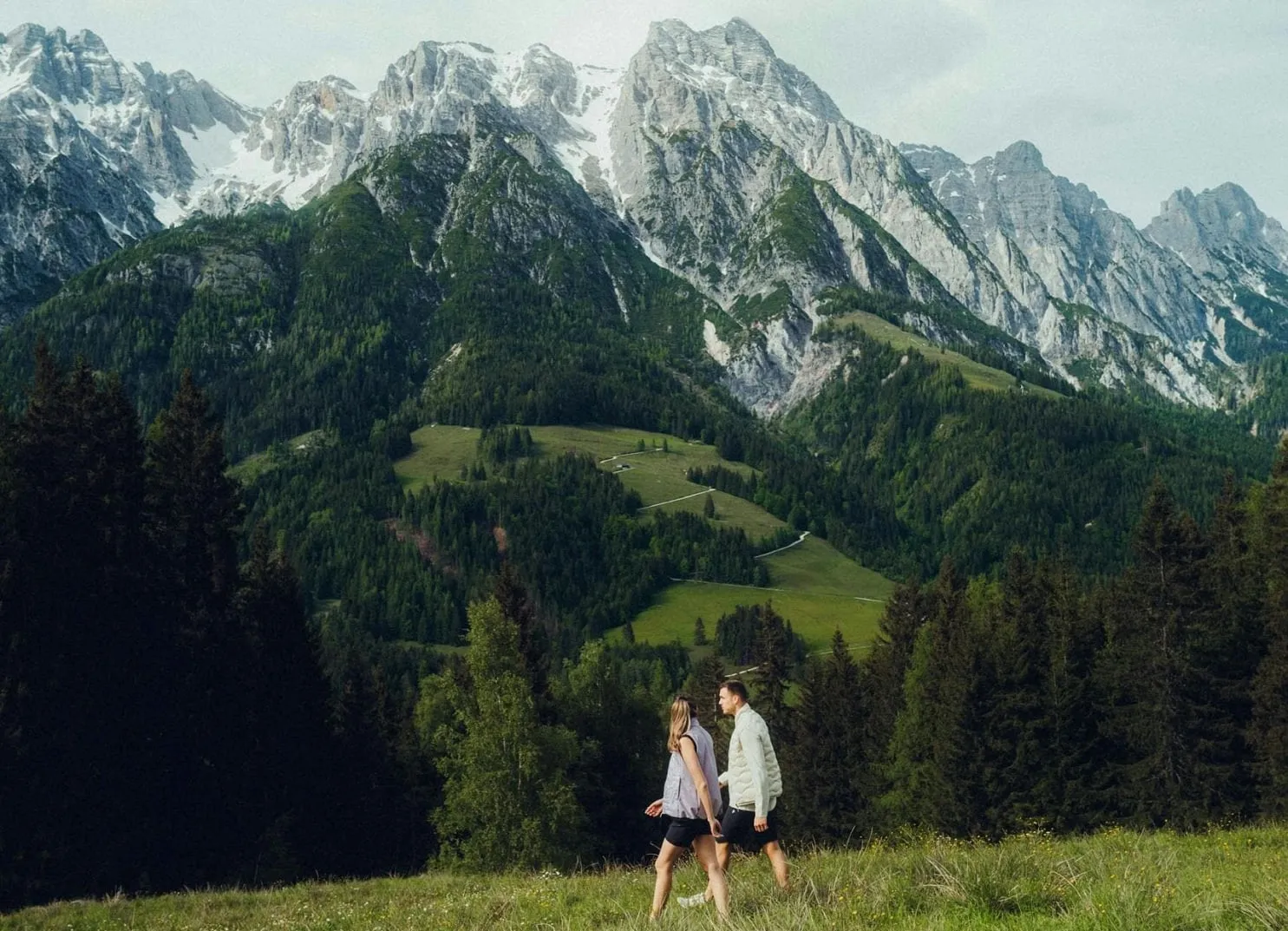Two people walking through a green field with mountains in the background