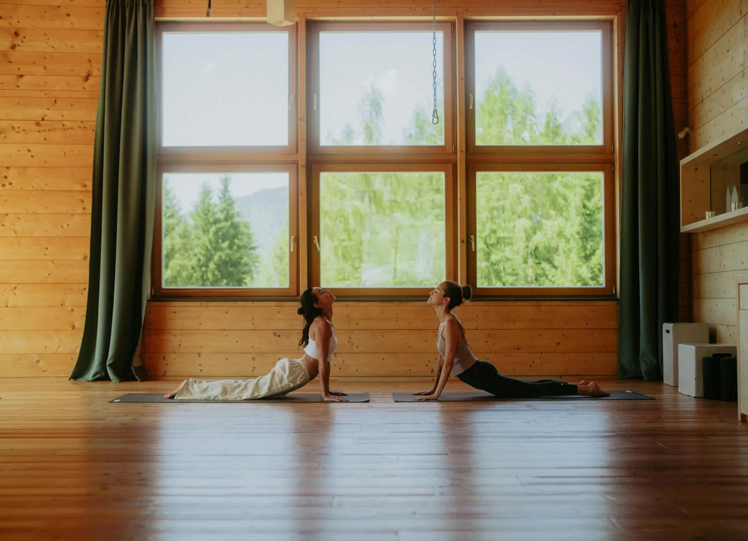 Two women practicing yoga in a wooden room with large windows and greenery outside.