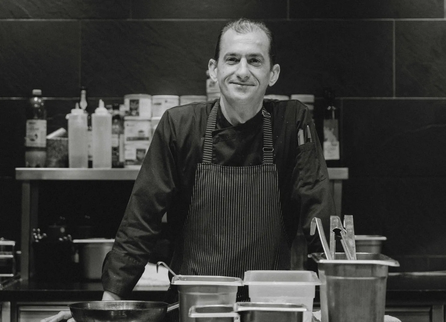 Chef in black apron smiling in a kitchen with pots and utensils.