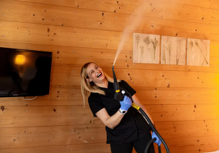 Woman with steam cleaner in wooden room, smiling in front of TV.