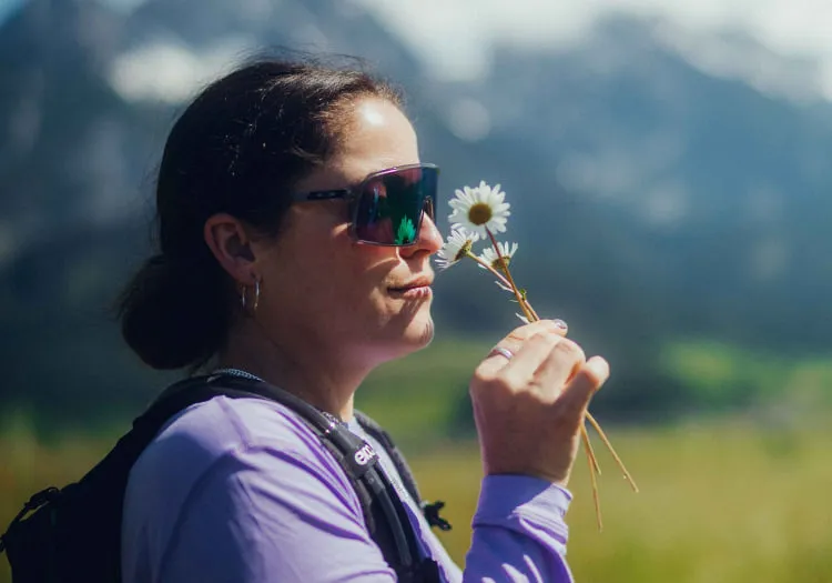 Woman in purple shirt holds flower, with mountains in background