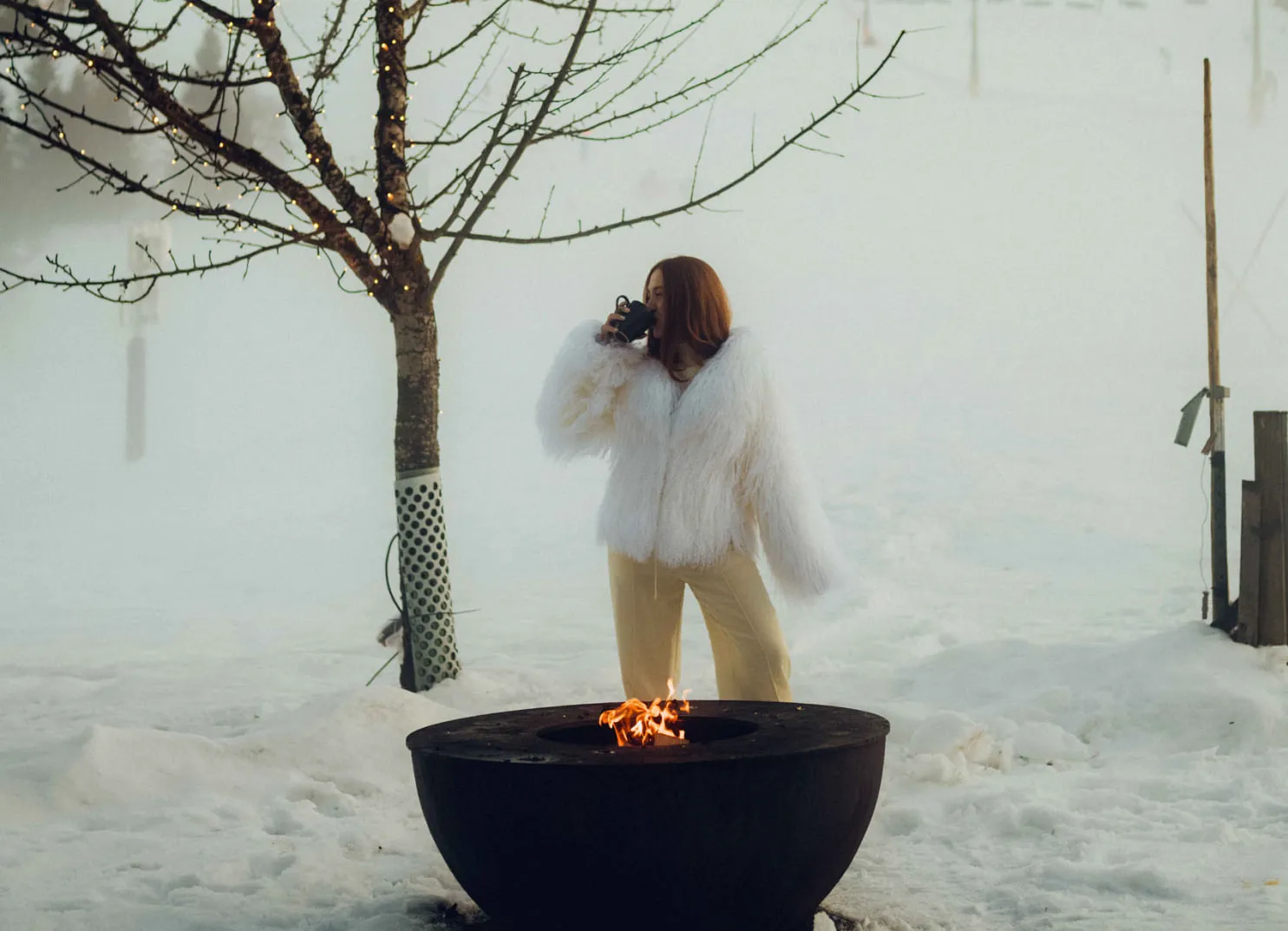 Frau in weißem Pelzmantel steht am Feuer im Schnee, Holzhotel Forsthofalm, Leogang