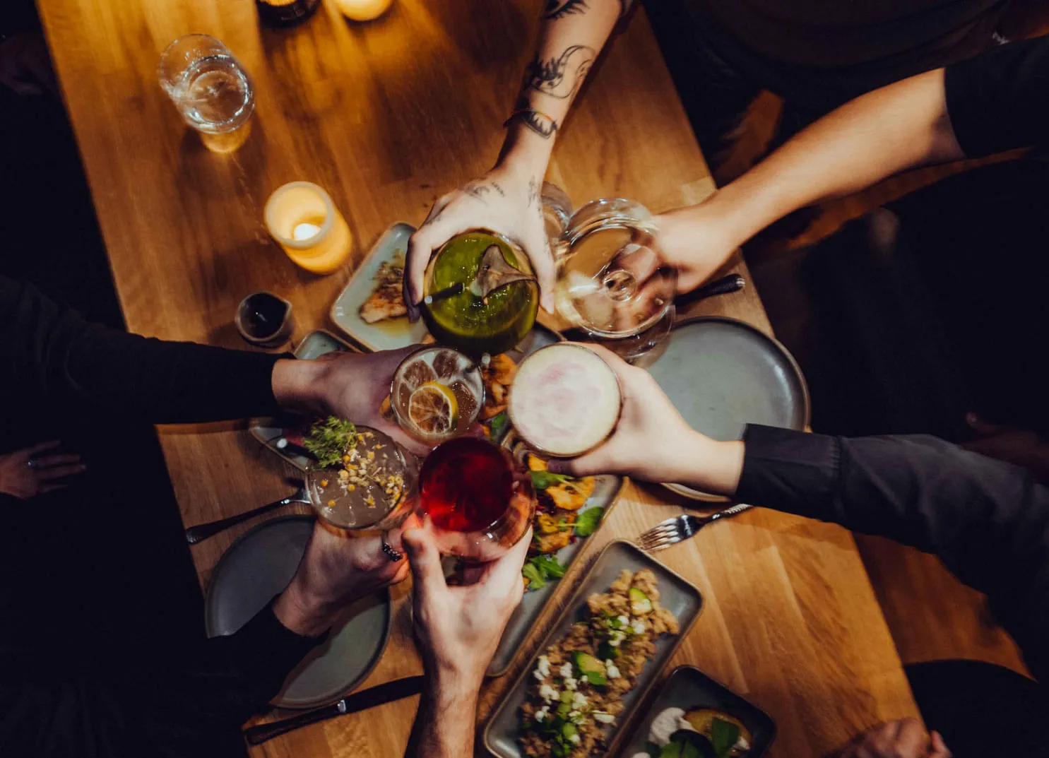 Mehrere Hände heben Gläser und Cocktails über einem Holztisch mit Essen.