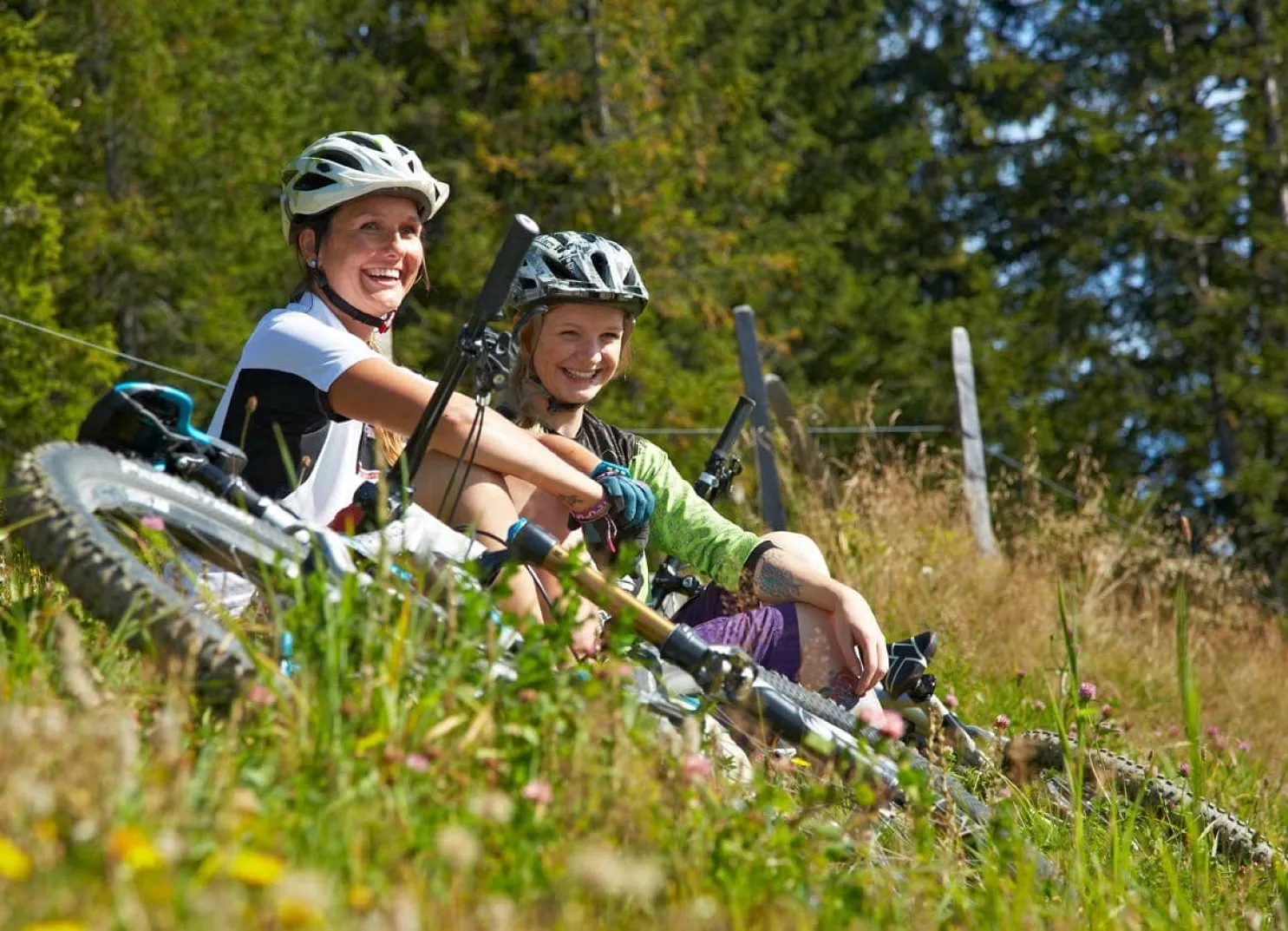Two smiling cyclists resting on a grassy hill with bicycles in a forested area