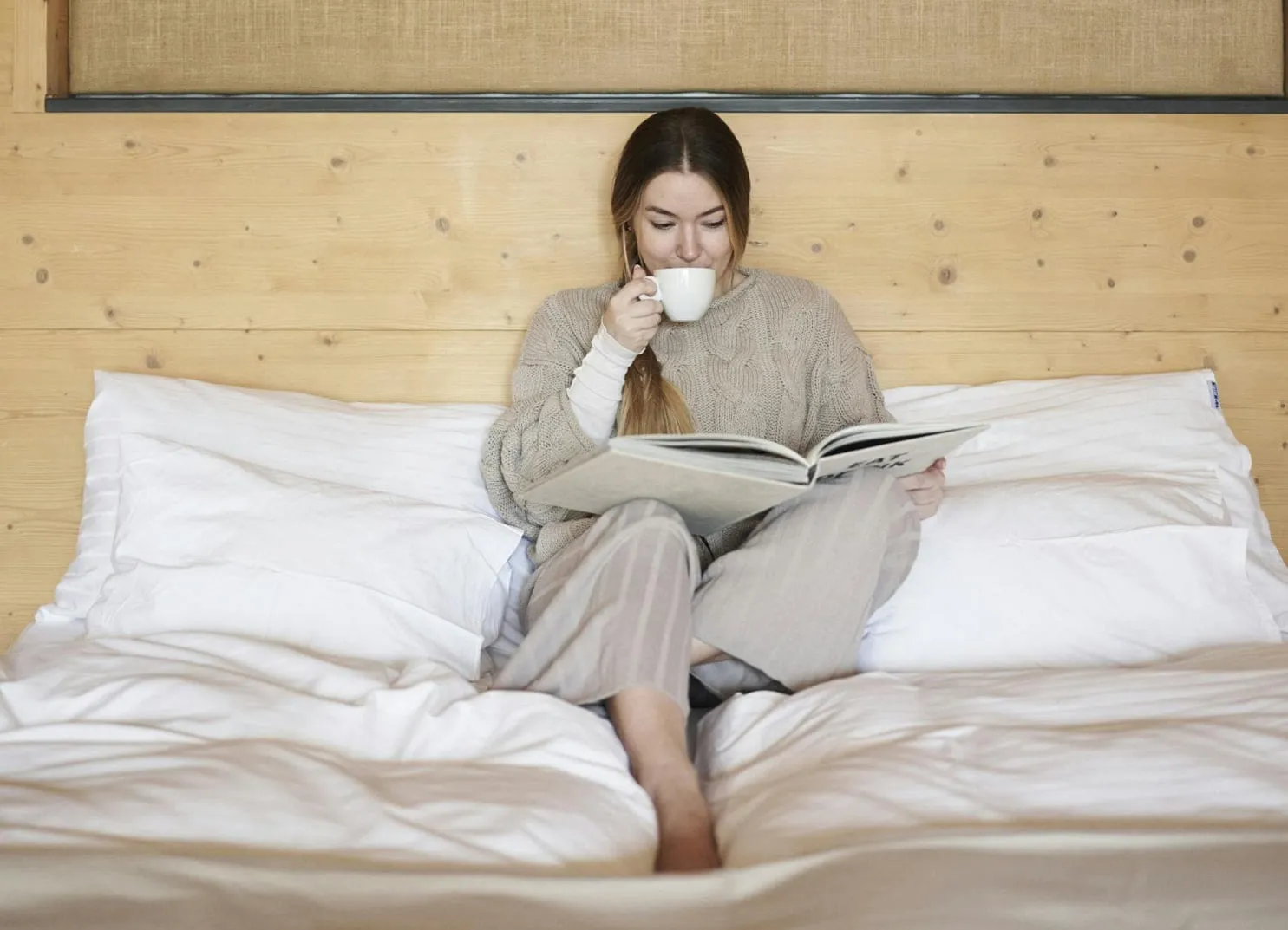Woman sitting on bed, reading a book and holding a cup.