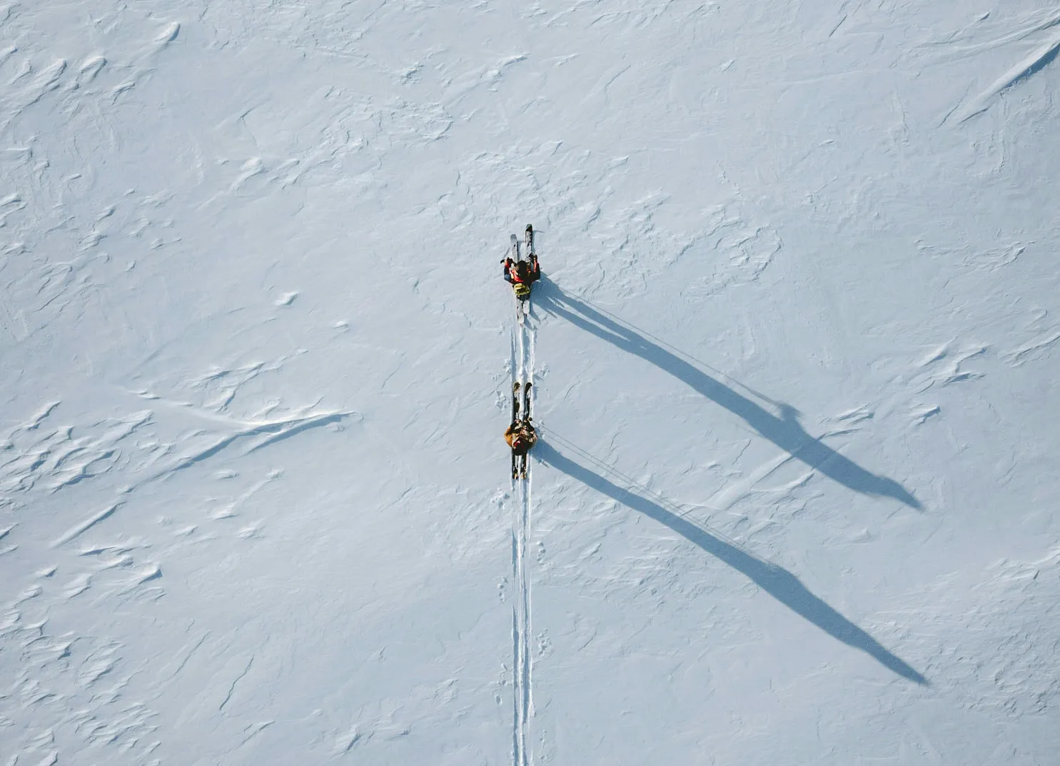 Zwei Skifahrer, die durch eine schneebedeckte Landschaft fahren und lange Schatten werfen.