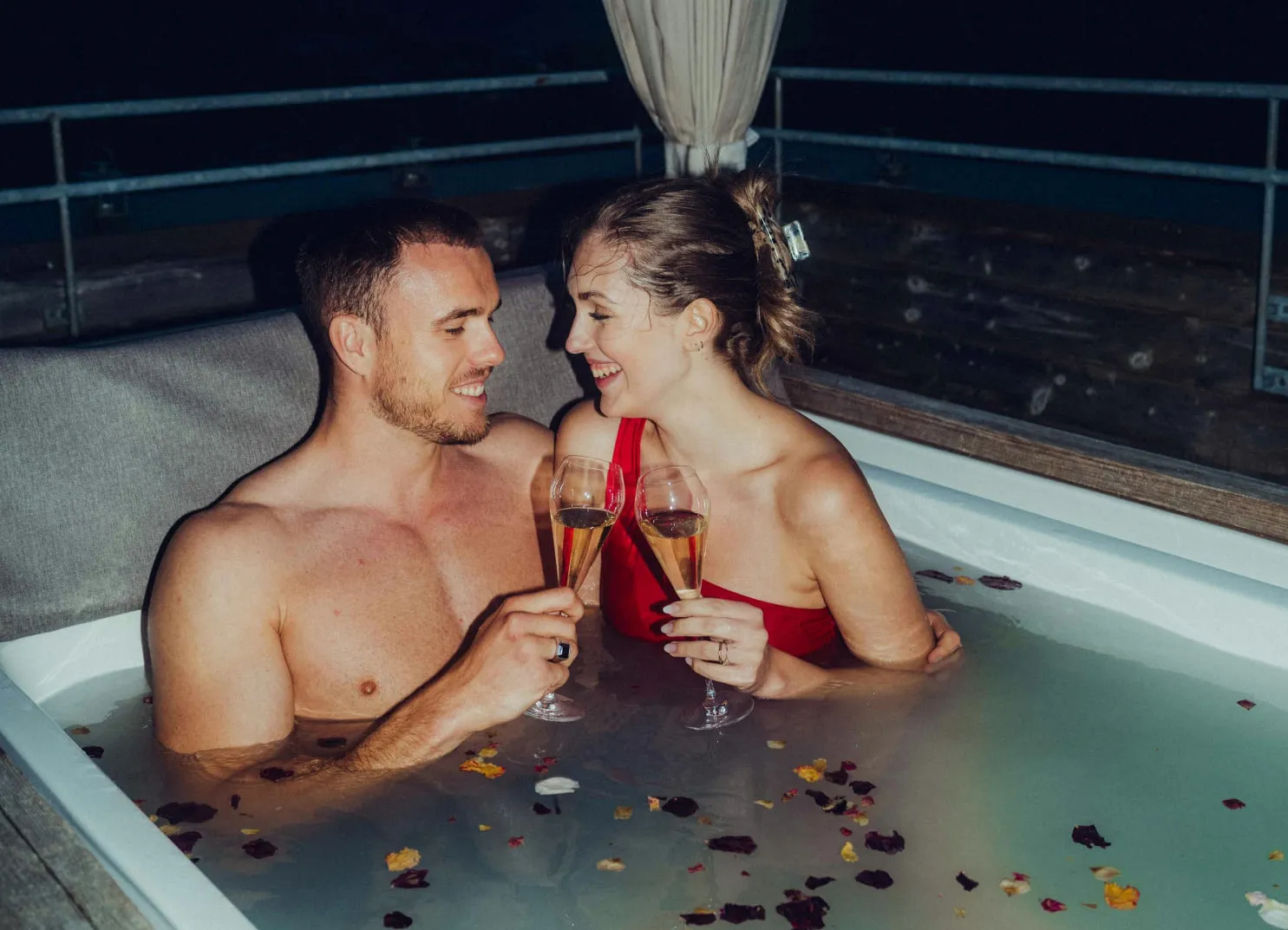 Couple in a hot tub at night, holding champagne glasses and surrounded by flower petals.