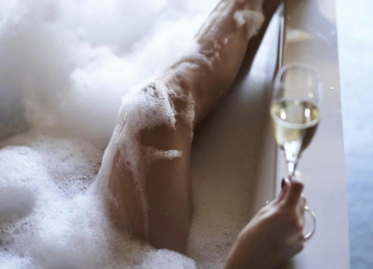 A hand holding a glass of champagne next to a leg in a bubble bath