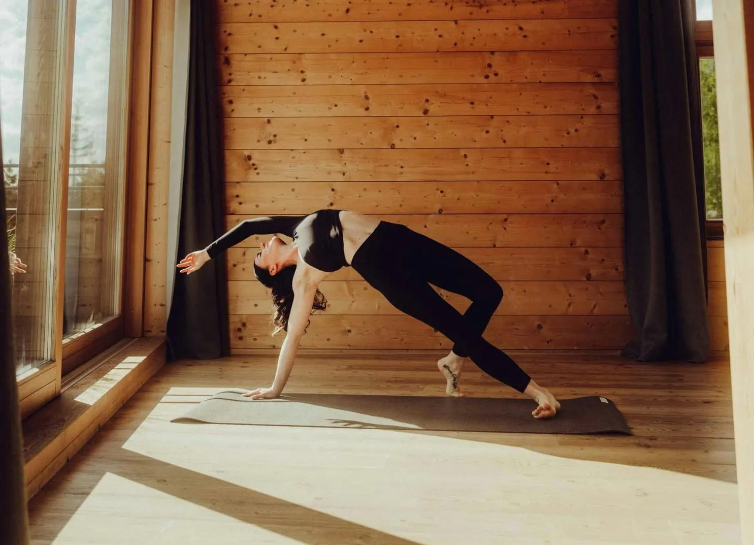 A woman performing a yoga pose in a wooden interior with large windows