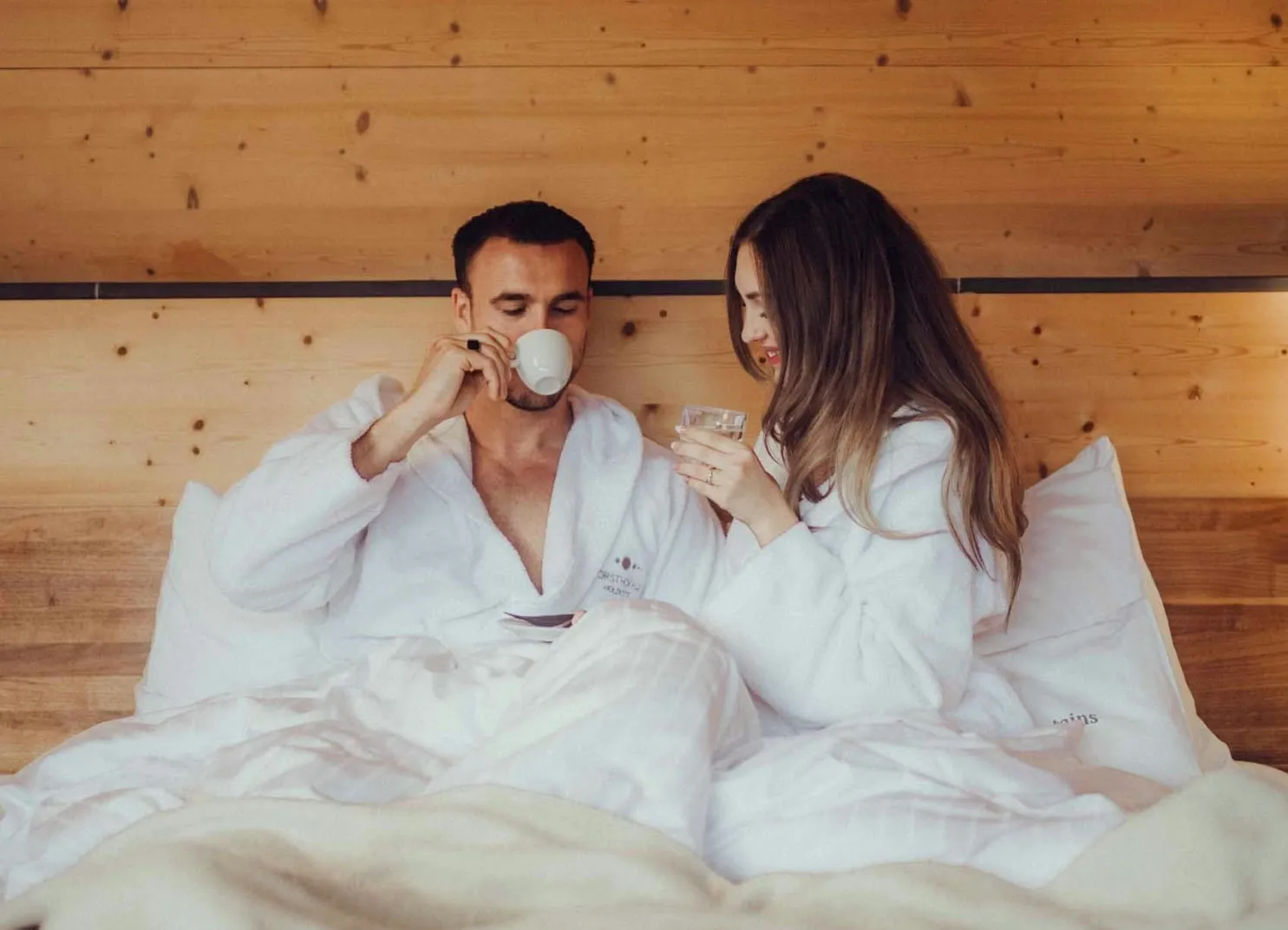 Paar in weißen Bademänteln entspannt im Bett mit Getränken