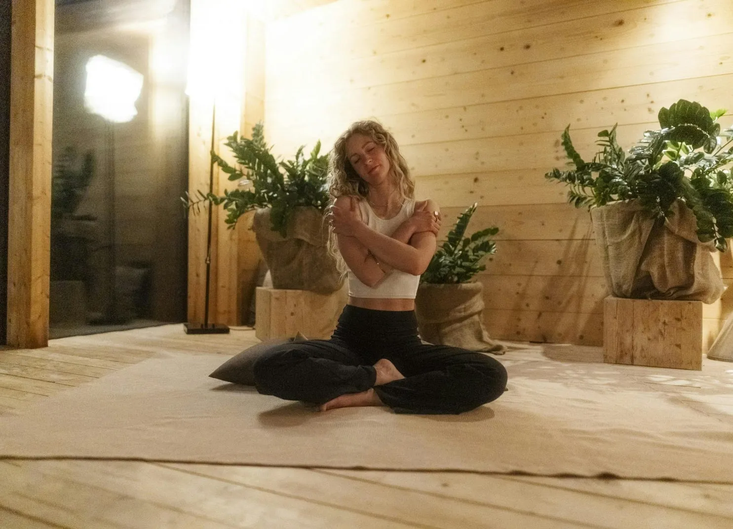 Frau, die im Lotussitz auf einem Teppich sitzt, umgeben von Pflanzen.