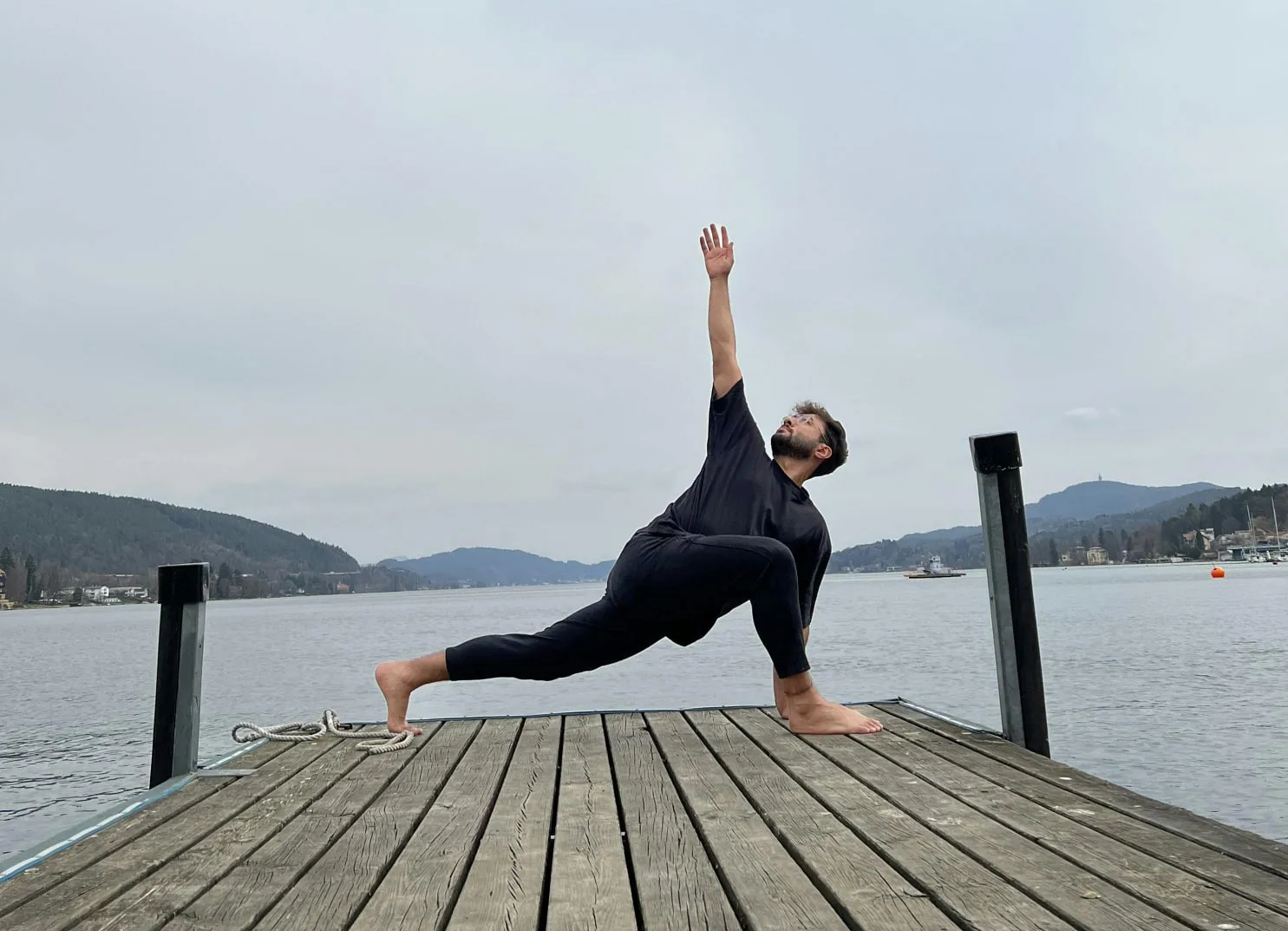 Person praktiziert Yoga auf einem Steg am See mit Bergen im Hintergrund