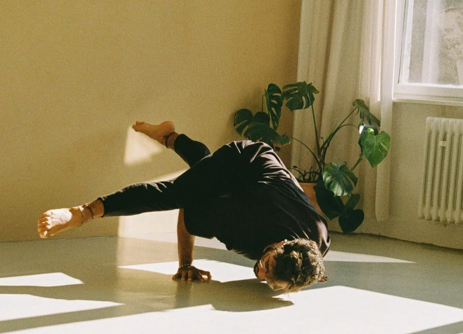 Ein Tänzer führt eine Bewegung auf einem hellen Boden nahe einem Fenster mit Pflanzen aus.