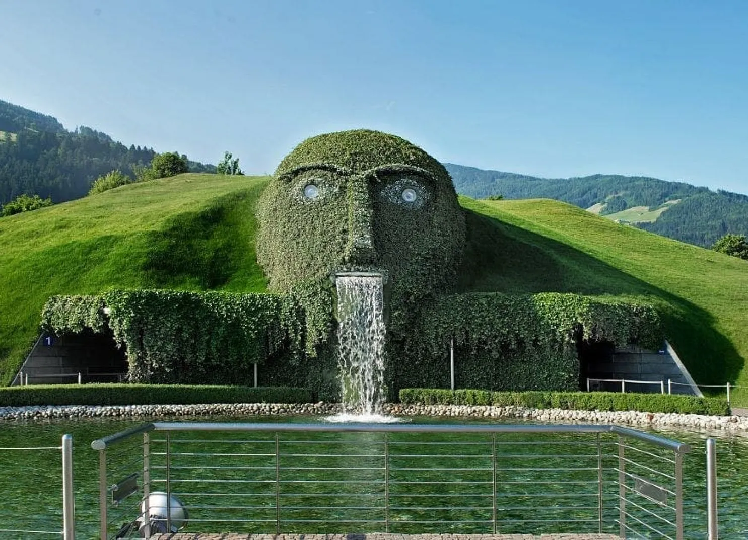 Ein großer grüner Hügel in Gesichtsform mit einem Wasserfall und einem See davor.