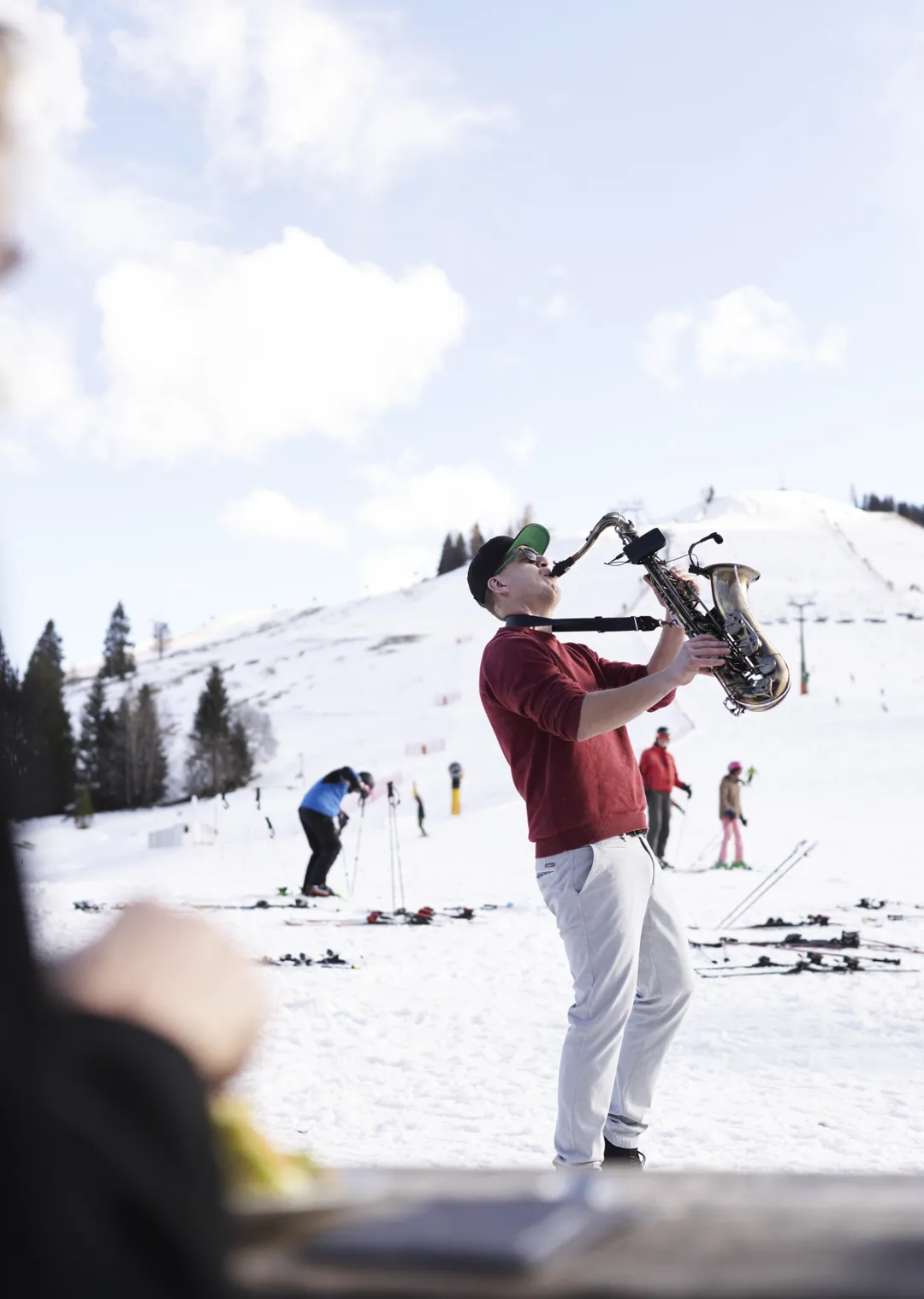 Mann spielt Saxophon auf einem schneebedeckten Hang, andere Skifahrer im Hintergrund