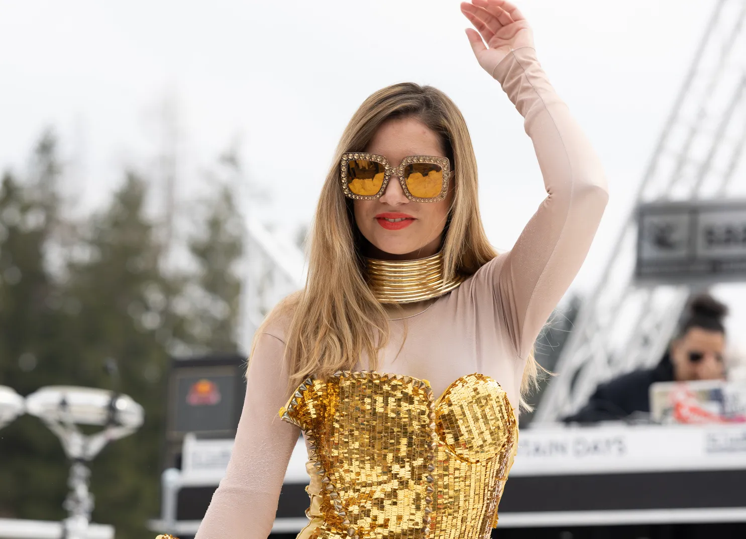 Person in goldenem Outfit und Sonnenbrille winkt, Hintergrund einer Veranstaltung im Freien