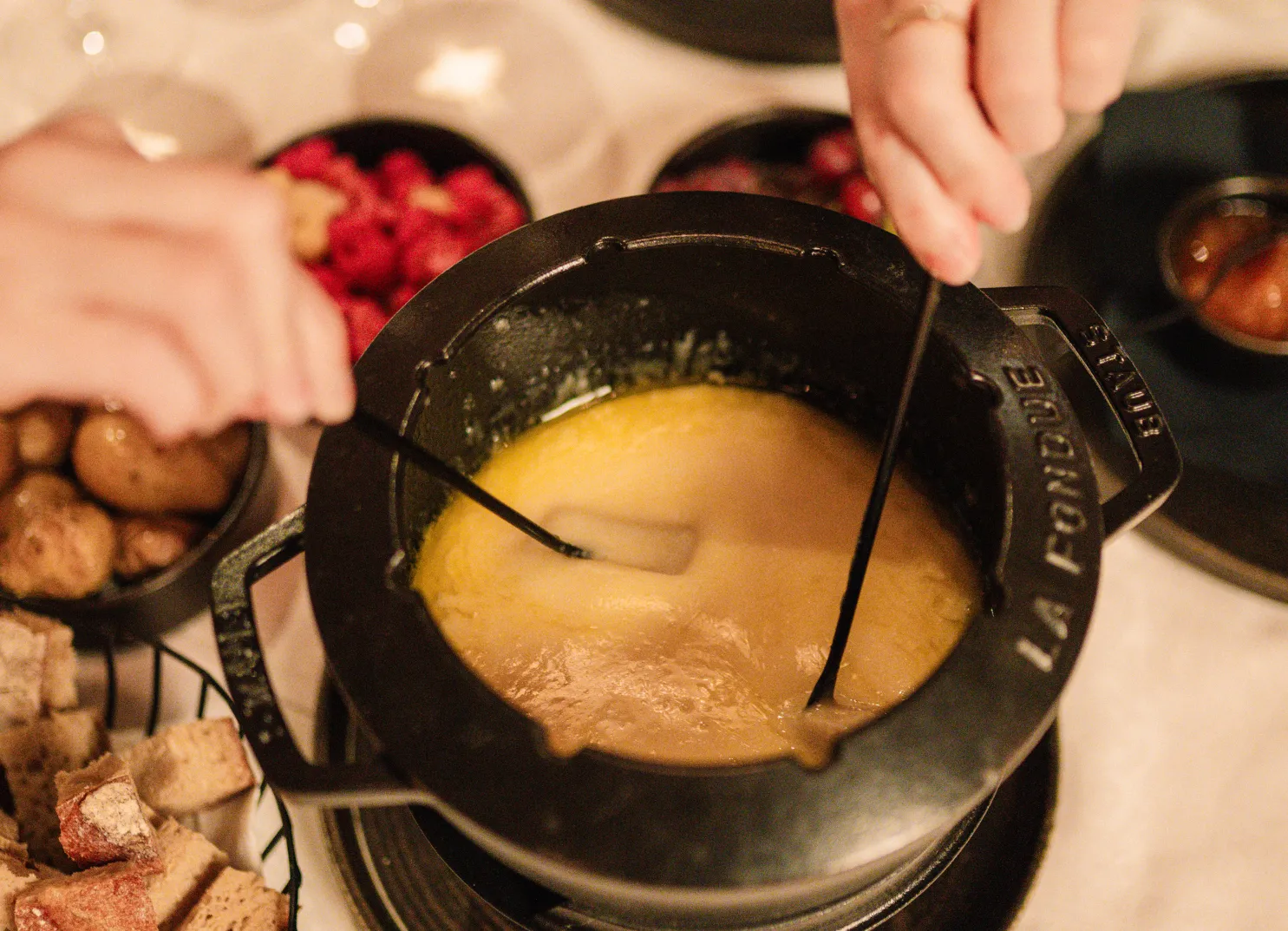 Fondue-Topf mit geschmolzenem Käse und Händen, die umrühren, umgeben von Snacks