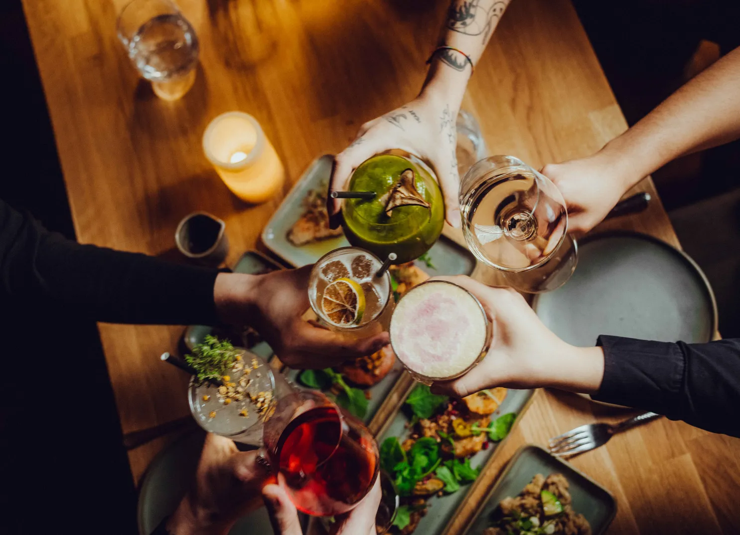 Hands holding cocktails above a table with food.
